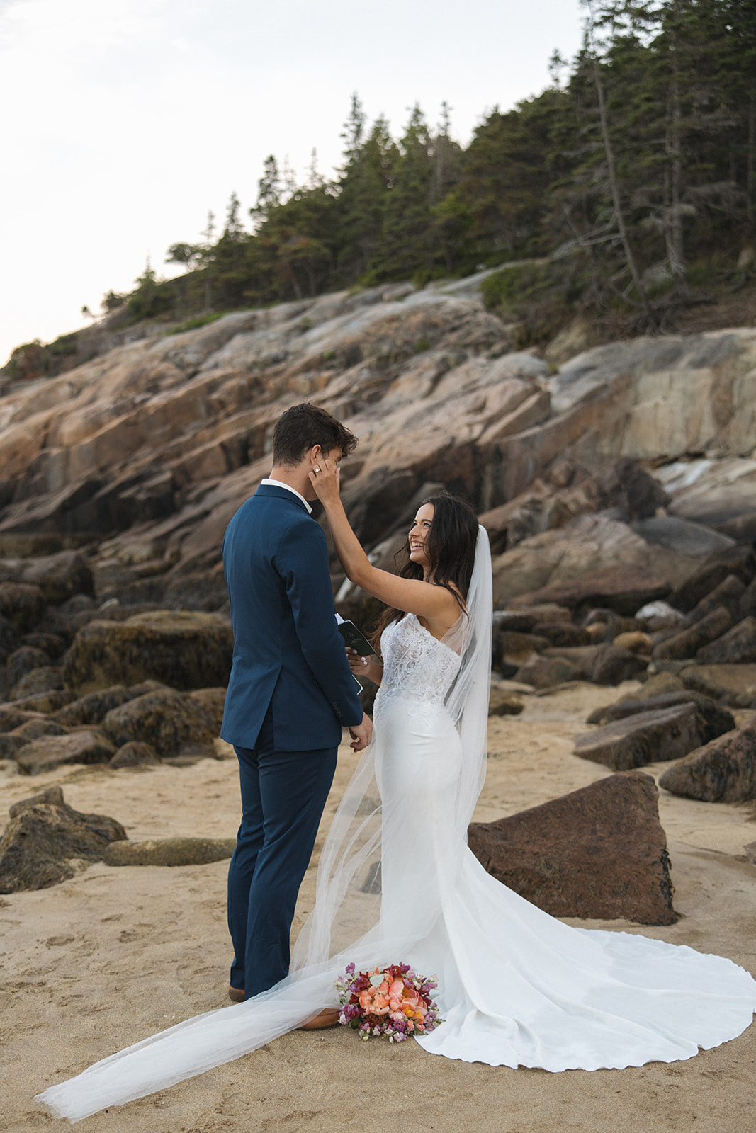 Bride wiping tear from grooms cheek while saying vows on the beach in Acadia National Park
