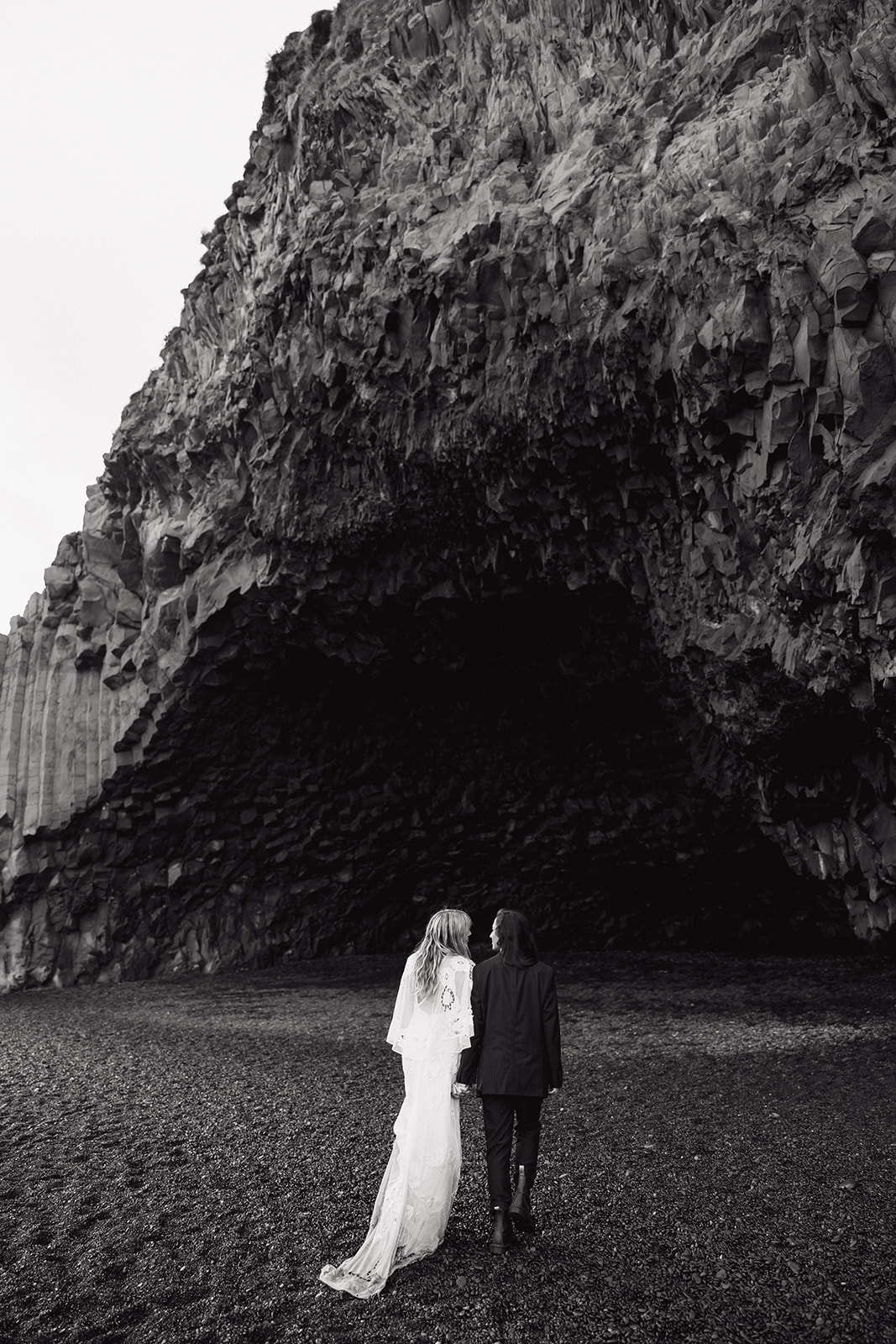 same sex couple walk towards a icelandic cave on the black sand beach