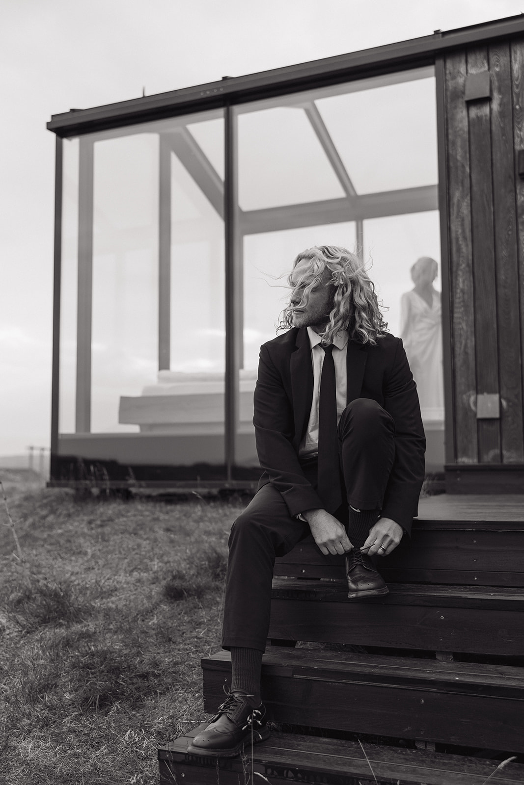 groom sits on stairs of glass house while his bride is getting dressed