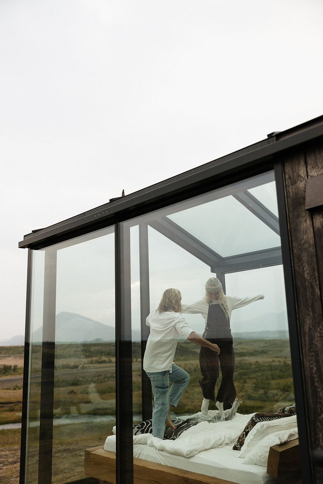 casual portraits of a bride and groom jumping on the bed in their glass house while in iceland