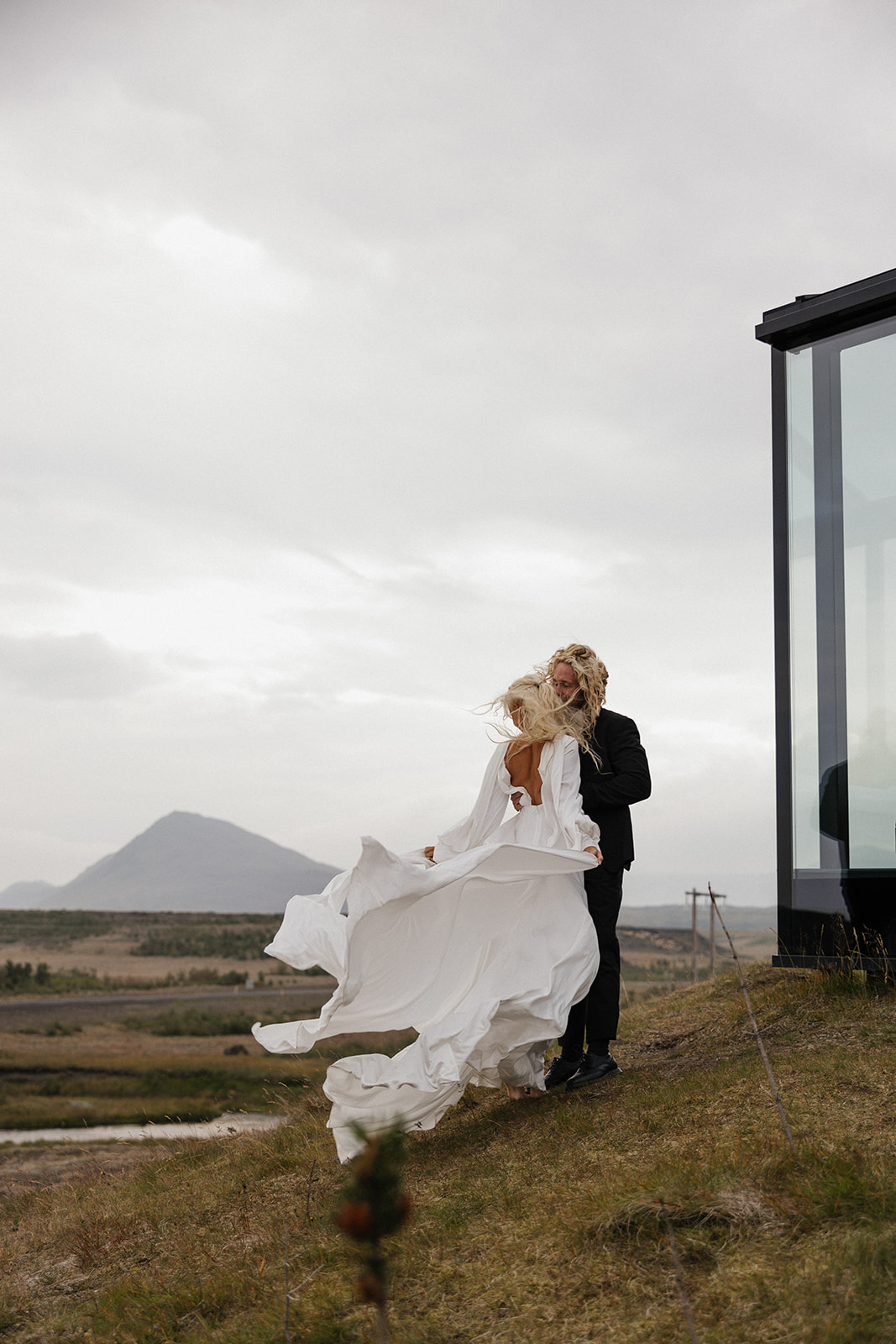 bride's white dress flows in the wind as she kisses the groom outside of glass house