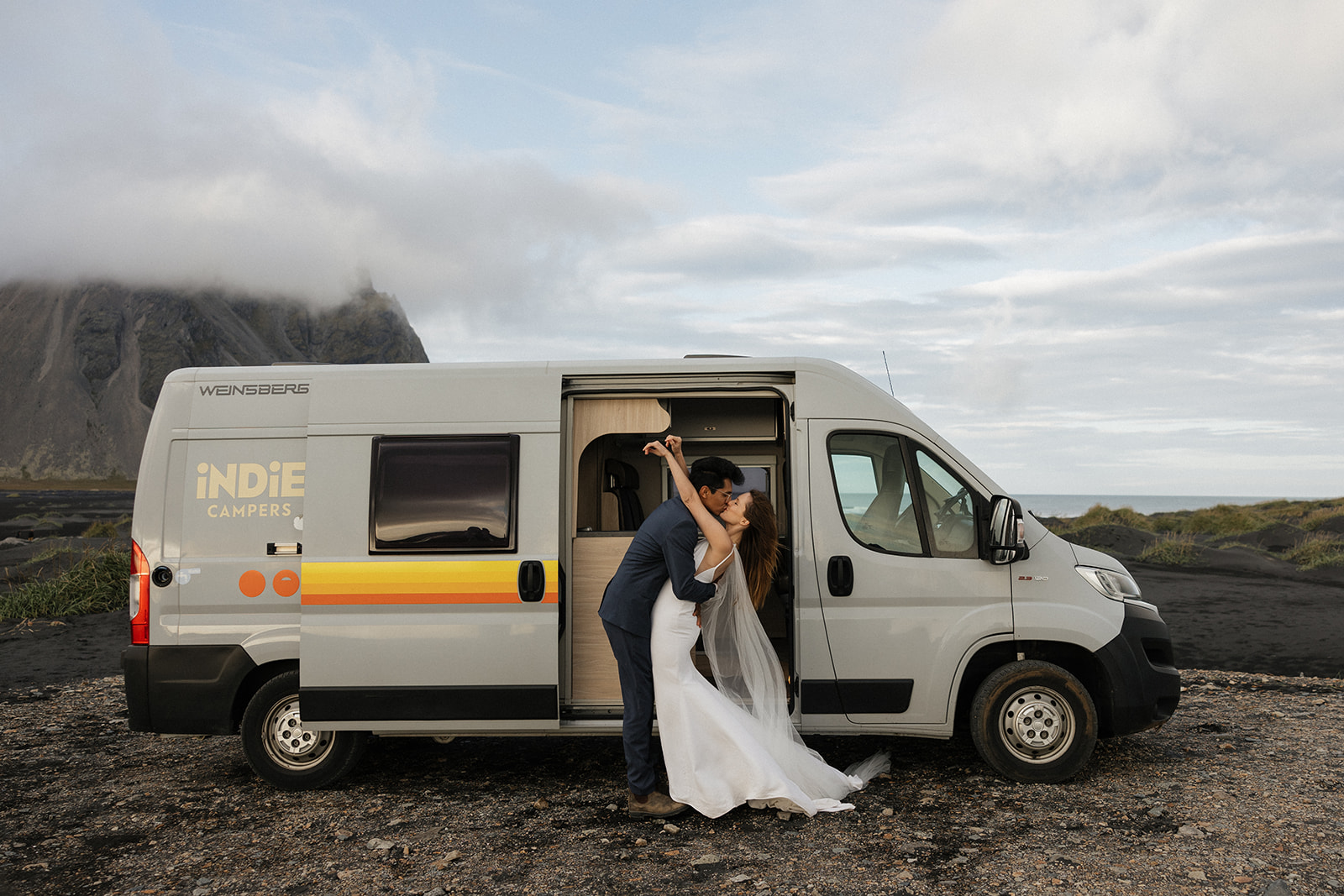 bride and groom kiss in front of their tan indi camper van while in iceland