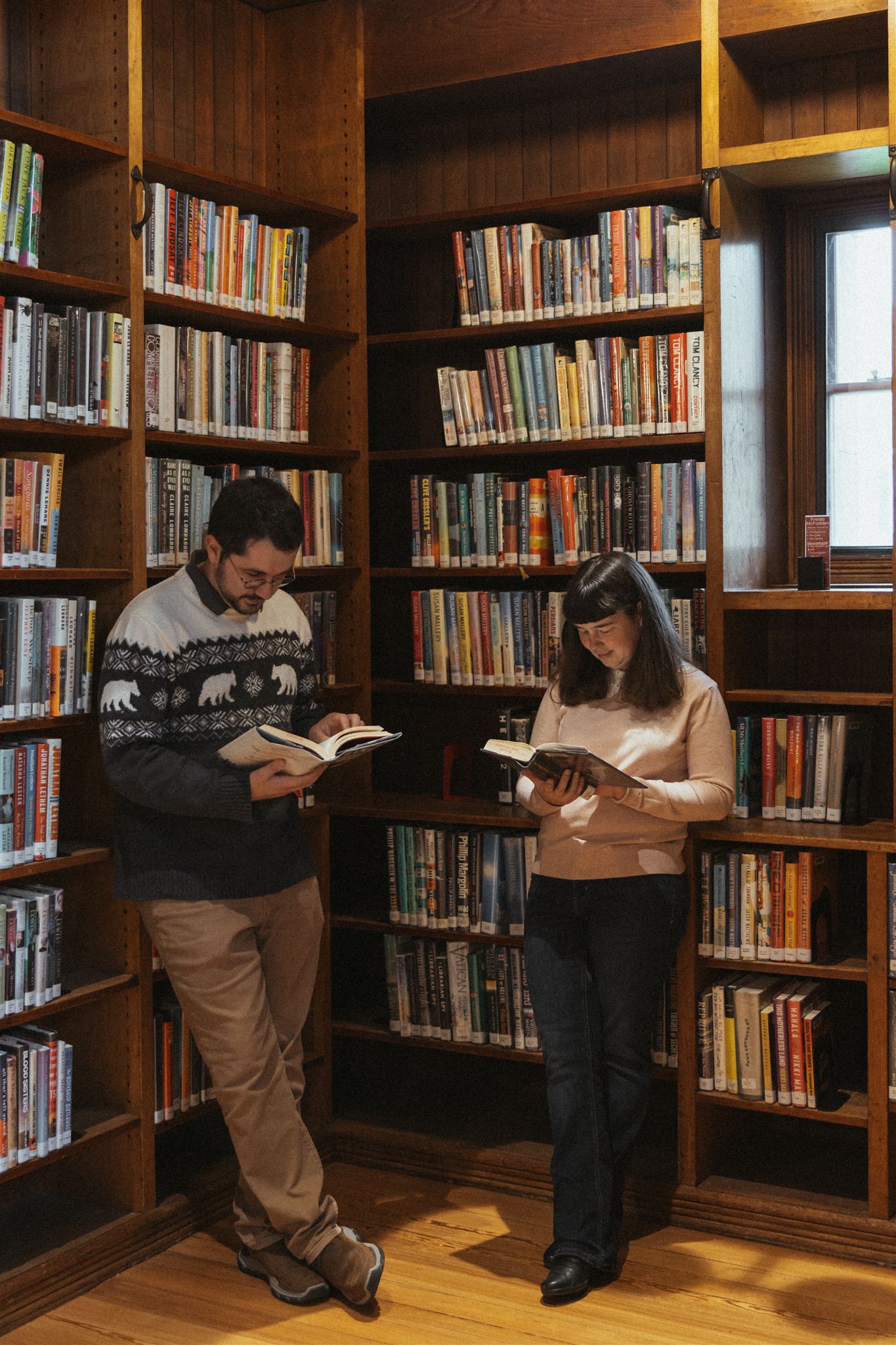 couple lean against the shelf reading books during their fun, cozy, and quiet engagement session