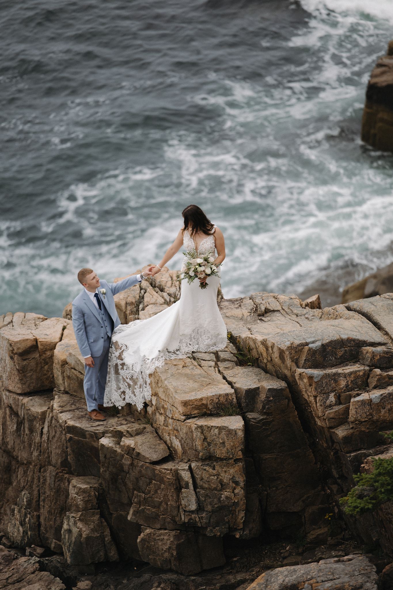 bride and groom hold hands and climb rocks together on their otter point acadia wedding day